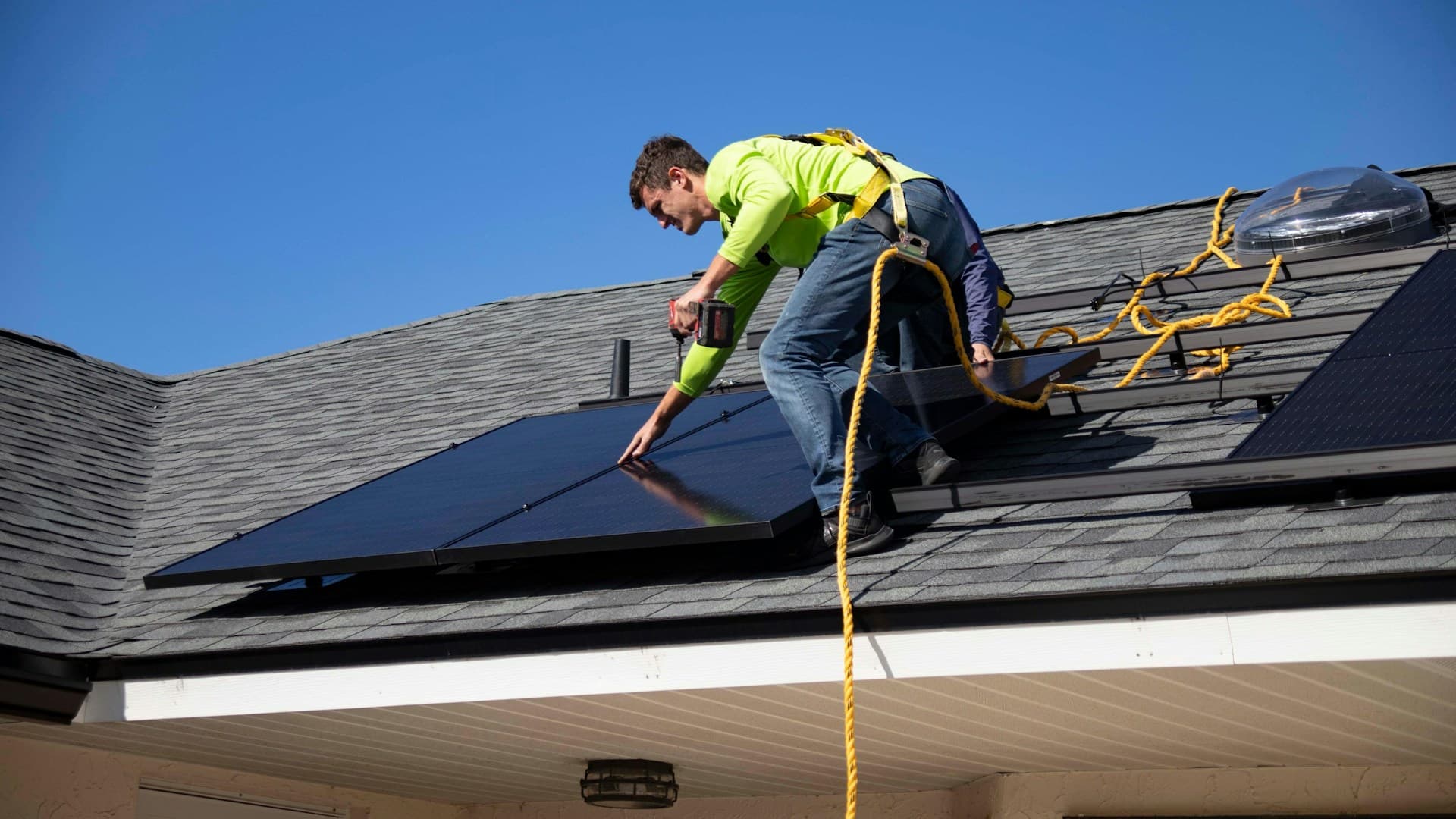 A tradie in hi-vis and a harness installing solar panels on a roof against the sky