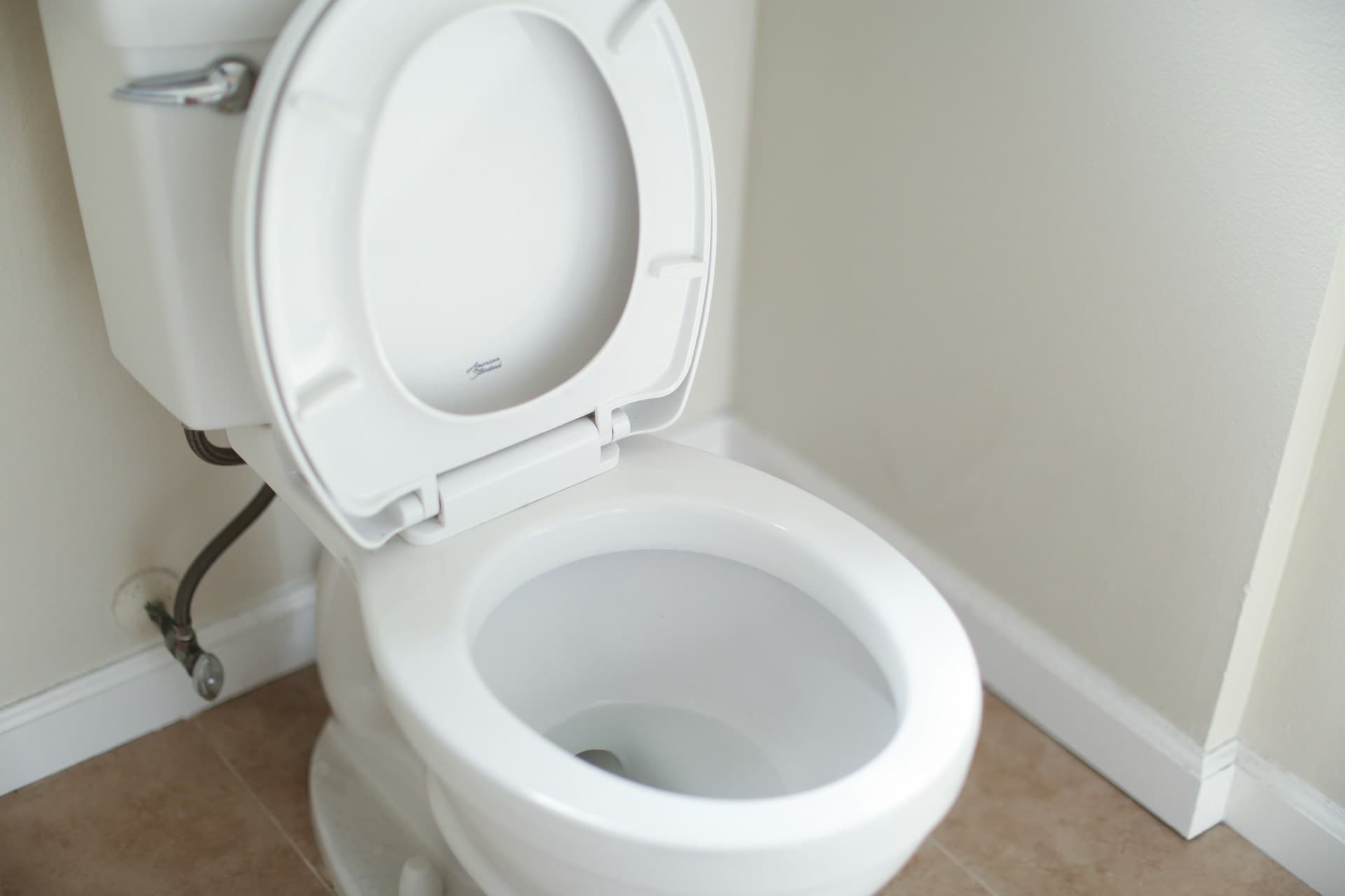 White ceramic toilet with lid open and exposed water supply line on tiled floor